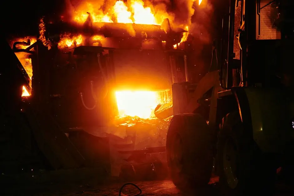 A dramatic industrial fire with flames engulfing machinery in a nighttime scene.